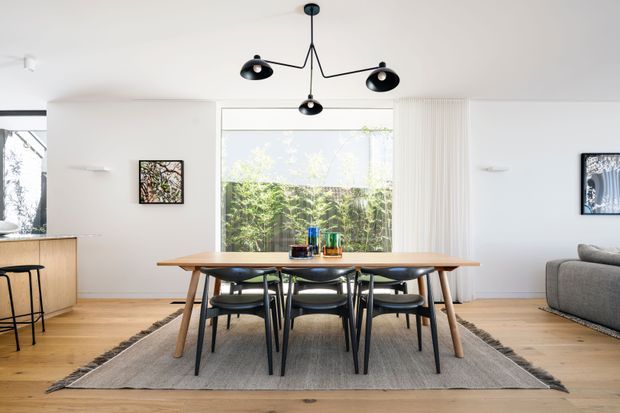 Bright, open-plan contemporary dining area with a large timber dining table and black dining chairs centred on a neutral rug. White walls, pale timber flooring, expansive glazing with vertical blinds, minimalist wall art, and a sculptural black multi-arm pendant create a clean, refined interior. A partial kitchen is visible to the left and a grey sofa to the right.