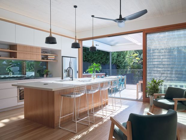 Bright open-plan kitchen and living area with a large timber island, white stone benchtop, integrated cabinetry, pendant lighting, and bar seating. Timber-framed sliding doors open to a covered outdoor deck, while a glazed wall and comfortable armchairs create a connection to the garden. The ceiling fan, warm timber finishes, and abundant daylight give the space a relaxed contemporary feel.