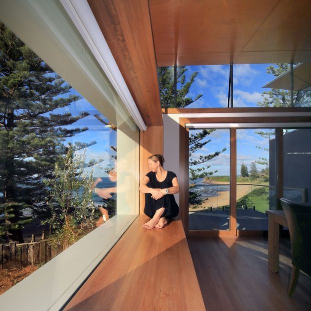 A bright contemporary interior corner with extensive floor-to-ceiling glazing overlooking mature trees and a coastal landscape. Warm timber-lined ceilings, walls, and a deep timber window seat contrast with white trim and dark-framed glass doors. A person sits on the built-in window seat, and part of a dining table and chair is visible at right.