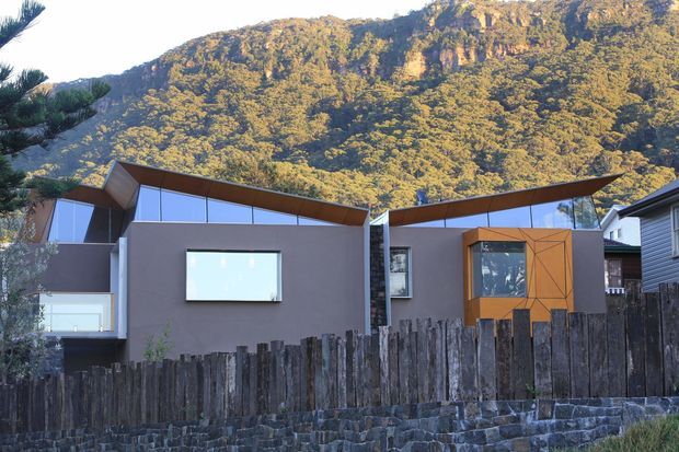 Exterior view of a contemporary multi-level residential building set against a steep, heavily vegetated rocky mountain backdrop. The facade combines dark rendered walls, extensive reflective glazing, angular projecting roof planes, warm timber soffits and a geometric timber-clad projecting bay. A rustic vertical timber fence and dark stone retaining wall occupy the foreground.