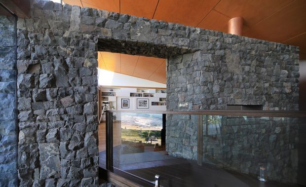 Interior architectural view dominated by a rugged dark grey natural-stone wall with a large rectangular opening and integrated dark-framed glass balustrade. Warm timber ceiling panels and a glimpse of a bright adjacent living area with artwork and shelving are visible beyond the opening. The composition emphasizes heavy masonry, strong geometric openings, and a dramatic warm-versus-cool material contrast.