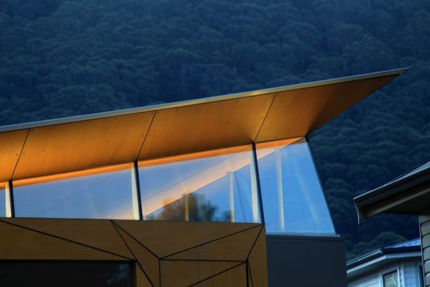 Close-up exterior architectural view of a sharply angular building with a projecting folded roof or canopy clad in warm timber, tall reflective glazing, and dark structural framing. A densely forested hillside forms the background, while part of an adjacent building is visible at the right edge. The composition emphasizes dramatic roof geometry, faceted planes, and the contrast between amber timber, blue glass, and dark cladding.