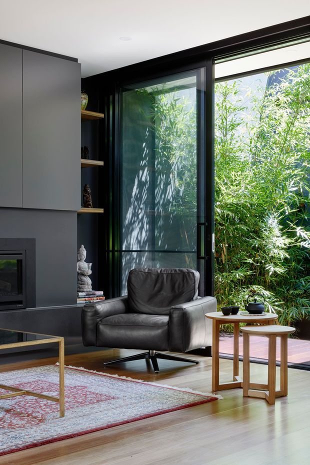 A compact, refined living room with a charcoal feature wall and built-in cabinetry surrounding a fireplace, open shelving, and a black-framed glazed door and window overlooking dense bamboo planting. The room includes a dark leather lounge chair, nested timber side tables, a slim metal-framed coffee table, a patterned area rug, and pale timber flooring. The overall look is contemporary with strong dark framing, natural light, and indoor–outdoor connection.