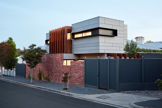 Street-front view of a contemporary multi-level residence with a boxy, flat-roofed form. The facade combines pale horizontal cladding panels, dark metal fencing and trim, warm vertical timber battens, and exposed red brick boundary walls. Narrow horizontal windows and projecting upper volumes create a strong modern composition.
