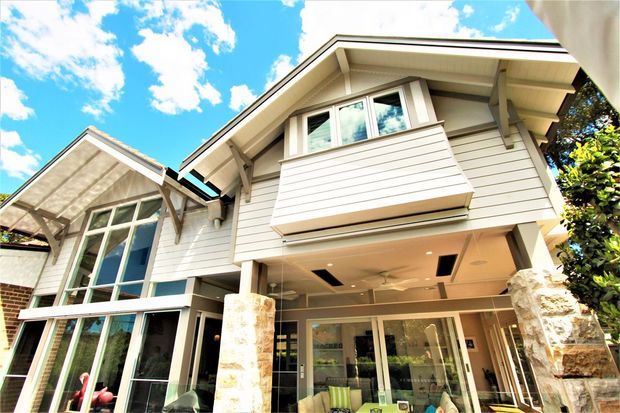 Upward-looking exterior view of a multi-level contemporary home with white horizontal weatherboard cladding, prominent gabled roof forms, exposed roof supports, large glazed openings, a projecting upper balcony, and substantial stone-clad columns at the ground-floor outdoor living area. Bright blue sky and surrounding greenery frame the facade.