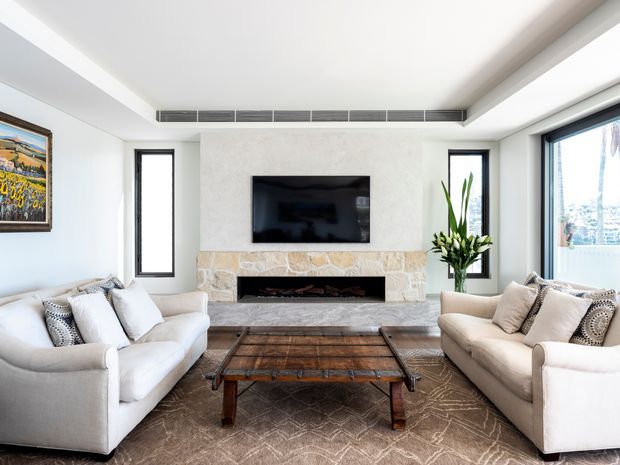 Bright contemporary living room with two large cream sofas facing a dark timber coffee table and a wall-mounted television above a natural stone fireplace. The room has white walls and ceiling, slim dark window frames, recessed linear ventilation, a large patterned neutral rug, and minimal decorative styling.