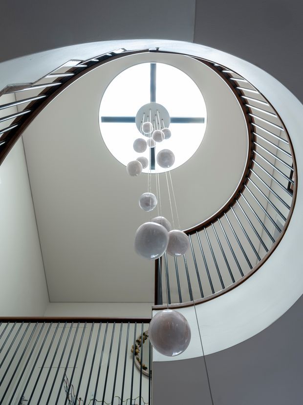 Upward-looking view through a sculptural curved stairwell with sweeping dark timber handrails, slim metal balustrades, smooth white walls, and a large circular skylight. A cascading cluster of translucent spherical pendant lights hangs through the stair void, creating a dramatic contemporary architectural interior.
