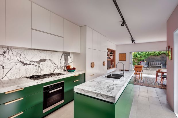 Contemporary open-plan kitchen with deep green lower cabinetry, white upper cabinetry, and a dramatic veined marble backsplash and island benchtop. The space includes an integrated cooktop and oven, undermount sink, timber-toned handles, track lighting, tiled flooring, and an open connection to a furnished outdoor courtyard or patio.