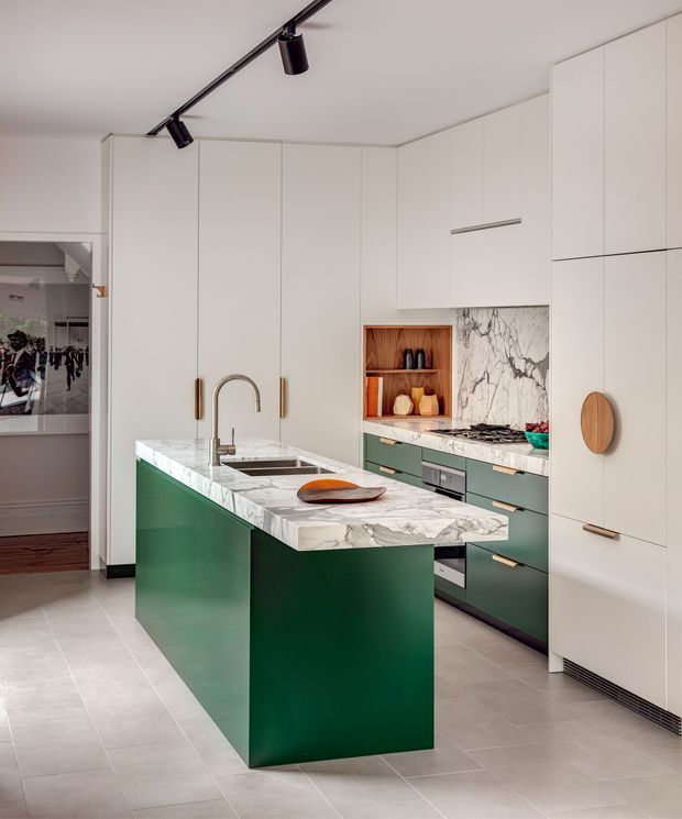 Contemporary kitchen featuring a large deep-green island with a white veined stone benchtop, integrated sink and curved brushed-metal tap. The surrounding cabinetry is predominantly white with slim timber handles, complemented by muted green lower cabinets, a marble-look splashback, and an open timber display niche. Pale large-format floor tiles and black track lighting complete the refined, high-contrast scheme.