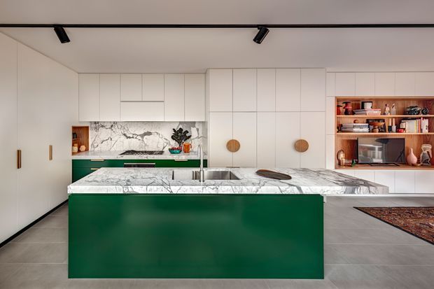 Contemporary open-plan kitchen dominated by a large marble island with a deep green waterfall-style base. Full-height pale cabinetry spans the back wall, with integrated appliances, a matching marble splashback, timber-toned open shelving, and warm brass or bronze handles. A tiled grey floor, recessed black track lighting, and minimal decor create a refined, modern interior.