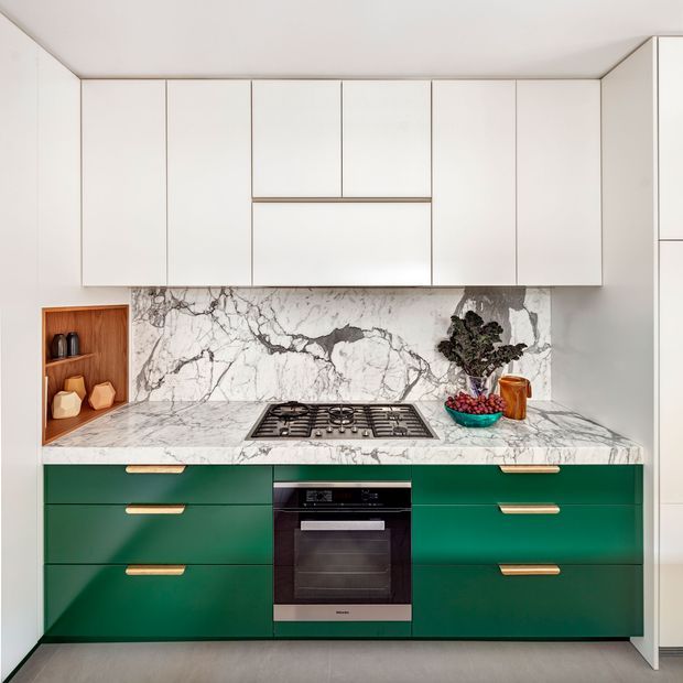 A compact contemporary kitchen viewed straight on, with floor-to-ceiling white upper cabinetry, deep green lower drawers, brass-toned handles, and a dramatic white marble-look stone benchtop and splashback. A gas cooktop and built-in oven are centred in the cabinetry, while an open timber-lined niche provides shelving at the left.