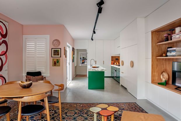 Open-plan contemporary kitchen and dining area with a compact timber dining table and chairs in the foreground, a green kitchen island, white full-height cabinetry, pale tiled flooring, and a built-in timber entertainment and shelving unit along the right wall. The room includes patterned artwork, a vintage-style rug, colourful nesting stools, and track lighting, creating a bright yet eclectic interior.