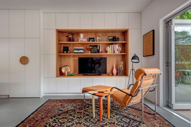 A refined living room with a built-in white storage wall framing a warm timber entertainment and display niche. The niche contains open shelving, books, decorative objects and a television. In front, a leather sling lounge chair and matching ottoman sit on a richly patterned Persian-style rug, with a slim black floor lamp beside them. Full-height glazed sliding doors open to an outdoor area, bringing in natural light. The space combines clean contemporary joinery with warm mid-century furnishings and earthy colour accents.