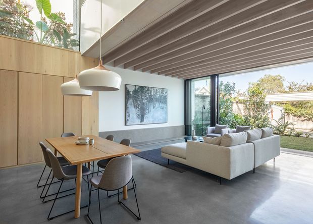 A bright, open-plan contemporary living and dining area with a timber dining table and upholstered dining chairs in the foreground, and a large light-coloured sectional sofa facing a low coffee table. Full-height glazing opens the living area to a leafy courtyard or garden. The room features a polished concrete floor, extensive warm timber wall cabinetry or panelling, a white wall, large pendant lights, and an exposed linear ceiling structure with visible beams or rafters.