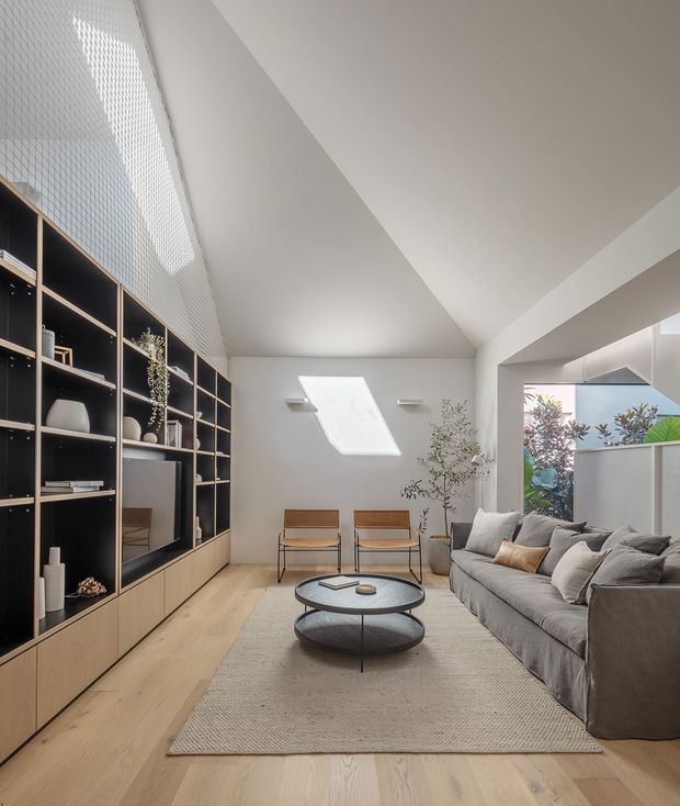 Bright contemporary living room with a large built-in floor-to-ceiling shelving and cabinetry wall in matte black and light timber, pale timber flooring, a large neutral rug, grey sectional sofa, round black coffee table, and two timber-framed lounge chairs. A sloped vaulted ceiling, skylight, clerestory-like glazing, and indoor planting create an airy, refined space.