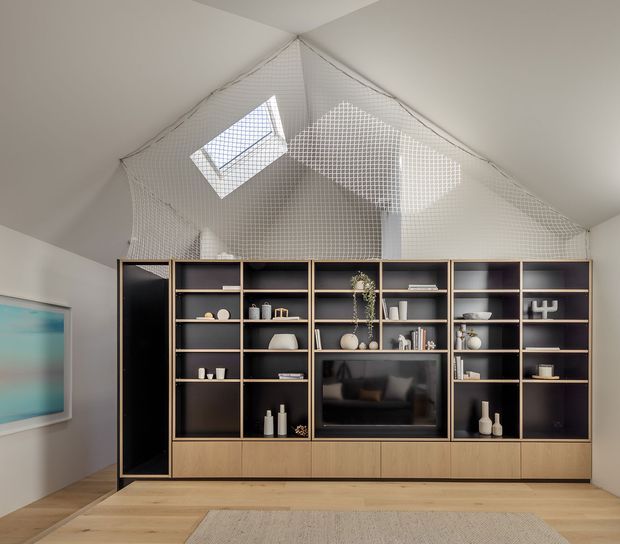 A refined living room with a large custom built-in media and display wall in dark cabinetry with warm timber edging and lower drawers. The cabinetry incorporates open shelving, decorative objects, books and a central television. The room has pale walls, light timber flooring, a large neutral rug, and a dramatic pitched ceiling with a high-level skylight and suspended safety netting beneath the ceiling void. A framed abstract artwork is visible on the left wall.