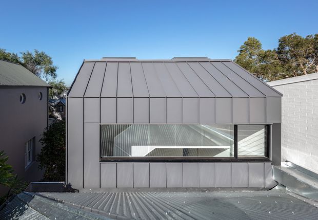 Exterior view of a compact contemporary rooftop structure with a steep folded metal roof and matching metal-clad walls. A large horizontal glazed opening reveals a bright interior with linear architectural detailing. The structure is set among neighbouring buildings and trees, with a corrugated metal roof in the foreground.
