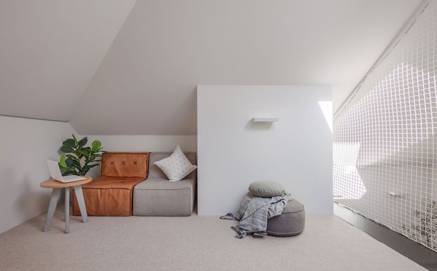 A bright, compact attic-style lounge beneath a sloping ceiling. The room features a small modular sofa with a warm tan leather seat and grey upholstered section, a round side table with a leafy plant, a low upholstered ottoman with a folded throw, and a large white tiled or gridded glazed opening along the right side. The palette is soft white, light grey, beige, and tan, creating a calm contemporary setting.