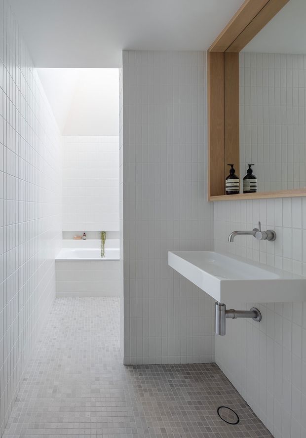 A bright, minimalist bathroom with white vertically gridded wall tiles, small pale mosaic floor tiles, a wall-mounted white trough basin with exposed chrome plumbing, and a recessed timber-framed mirror niche with built-in shelf and dark soap dispensers. A freestanding white bathtub is visible through the open bathing area, with a skylit or clerestory-like opening providing soft natural light.