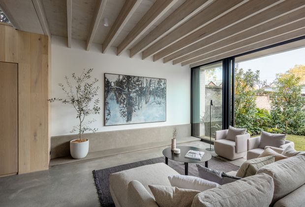 A refined contemporary living room with a large sectional sofa and two armchairs arranged around a round coffee table. The room features a light concrete floor, a long integrated concrete bench or ledge, pale timber wall paneling and exposed timber ceiling beams. Full-height black-framed glazing opens the room visually to a planted garden, while a large artwork and potted tree add restrained decorative elements.