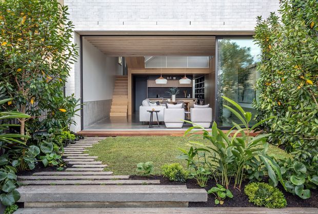 A landscaped courtyard garden leads to a contemporary home through a wide open glazed living area. The facade combines pale brickwork, expansive glass sliding doors, and warm timber-lined soffits and stairs. Inside, a furnished living and dining space with a dark kitchen or bar joinery wall is visible. Stepping pavers cross the lawn, framed by dense lush planting.
