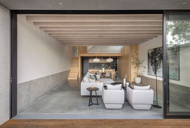 A contemporary open-plan living and dining space viewed through large dark-framed sliding glass doors. The room features a polished concrete floor, white plaster walls, a partial concrete wall finish, exposed light timber ceiling beams, timber cabinetry and stair elements, and a compact kitchen at the rear. White upholstered lounge chairs and a sofa sit around a small dark side table, with floor-to-ceiling glazing opening toward an outdoor courtyard or garden.