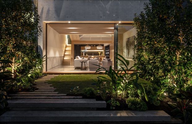 Night-time exterior view through a landscaped garden toward a contemporary home. Broad concrete steps and a narrow stepping-stone path lead to a fully glazed opening framed in dark metal, revealing a warmly lit living and dining space. The projecting opening has a timber-lined ceiling, while dense tropical planting and integrated landscape lighting surround the facade.