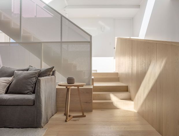 A bright, minimalist split-level living area with pale timber flooring, built-in timber stair and wall joinery, and a grey upholstered sofa in the foreground. A perforated translucent screen with slim framing separates the seating area from the stairs, while daylight enters from high-level glazing or a skylight. The palette is predominantly white, pale timber, and soft grey, creating a calm, airy interior.