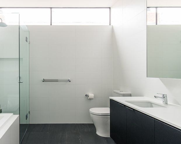 Bright contemporary bathroom with white tiled walls, a large frameless glass shower enclosure, wall-hung dark vanity cabinetry, white benchtop with integrated basin, toilet, chrome fixtures, and high-level windows providing natural light.