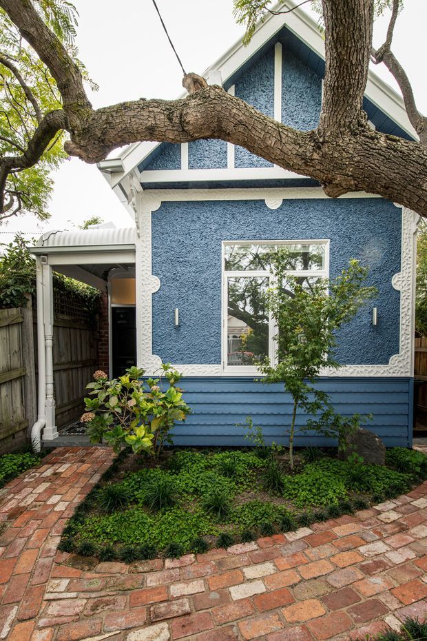Exterior view of a compact, heritage-style blue cottage or detached dwelling, framed by a large mature tree. The facade has textured blue stucco, white decorative trims, a steep gabled roof, white-framed window, and blue horizontal weatherboard cladding below. A covered side entry and brick-paved garden path sit among dense low planting and timber fencing.