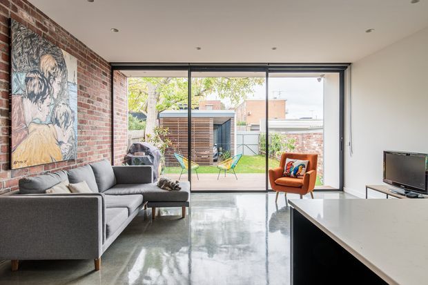 Bright contemporary living room with polished concrete flooring, exposed red-brick feature walls, a grey sectional sofa, a vivid orange armchair, and full-height sliding glass doors opening to a small landscaped courtyard. A minimalist white kitchen island is visible in the foreground, with an adjacent outdoor pavilion or studio beyond the garden.