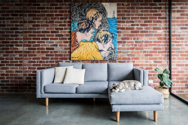 A stylish living room with an exposed red brick feature wall, polished grey concrete floor, and large black-framed glazing. A light grey sectional sofa with cream cushions and a small dog sits beneath colourful artwork, accompanied by a potted plant. The space has an urban loft-like character with strong natural light.