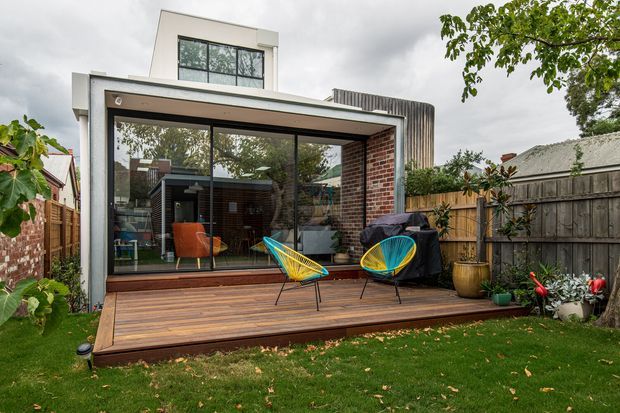 Rear exterior view of a contemporary two-storey residential extension with a box-like upper level, dark-framed floor-to-ceiling glazing, exposed red-brick wall, and a timber deck opening onto a suburban garden. Bright woven outdoor lounge chairs sit on the deck, with timber fencing and neighbouring residential structures surrounding the space.