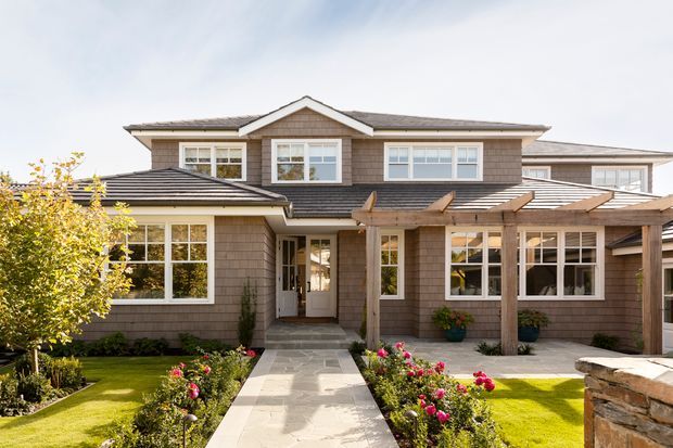 Front exterior view of a large two-storey suburban home with shingle-style cladding, multiple gabled roof sections, white-framed windows, a covered entry, and a timber pergola extending over the front walkway and patio. The landscaped frontage includes lawns, planting beds, rose bushes, and a stone boundary wall.