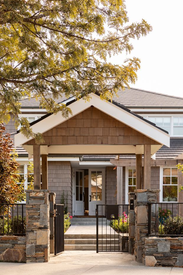 Front exterior view of a substantial residential entrance, framed by mature trees. A covered gabled porch with timber posts, a shingle-clad gable, stone masonry piers and low stone walls leads to a glazed entry door. The wider house has grey shingle or weatherboard-style cladding, light trim, and a broad hipped roof with dark grey tiles or shingles. A metal pedestrian gate, paved path, garden planting and black railings complete the approach.