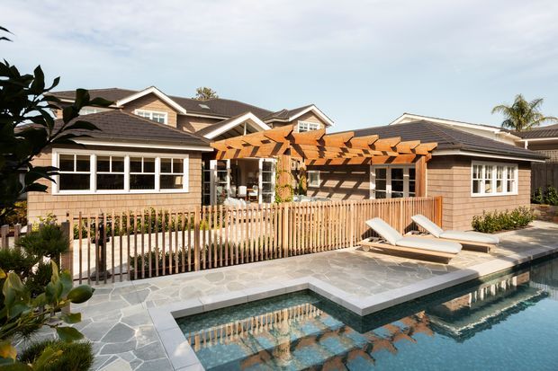 Exterior view of a large, low-rise residential property surrounding a swimming pool and paved terrace. The building has shingle-style wall cladding, dark tiled hip and gabled roofs, white-trimmed windows, and a timber pergola over a sheltered outdoor entertaining area. A timber picket fence, sun loungers, stone paving, and landscaped planting complete the poolside setting.