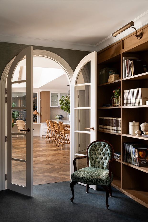 A refined study or library viewed through open arched glazed double doors. The foreground room has dark carpet, deep green textured wallpaper, extensive dark timber built-in bookshelves, a brass picture light, and a patterned upholstered armchair. Beyond the doors is a bright open-plan kitchen and dining area with parquet timber flooring, white cabinetry, a dining table, and woven timber dining chairs. The interior combines classical detailing such as arched doorways, panelled trim, and crown moulding with warm contemporary joinery.