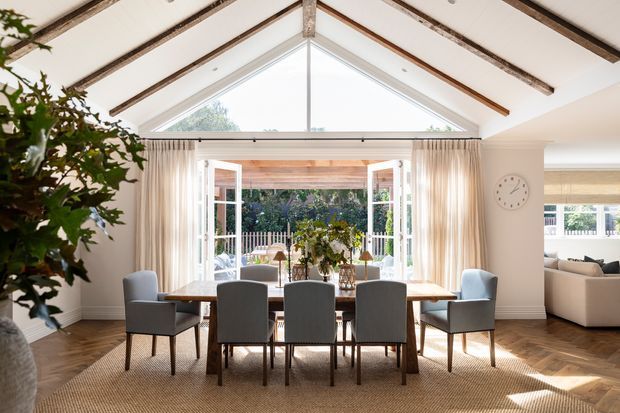 A bright, elegant dining room centred on a large rectangular timber dining table with upholstered dining chairs. The room has a high pitched, vaulted ceiling with exposed dark timber beams, white walls, herringbone timber flooring, and a large glazed gable with French doors opening to a leafy garden and outdoor seating. Full-height sheer curtains, a neutral rug, and recessed lighting create an airy, refined interior.