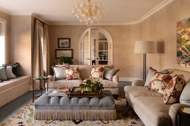 Elegant, warm living room with a symmetrical seating arrangement, including two sofas, a large fringed ottoman, and a window seat. The room features beige textured wallcovering, pale upholstered furniture with floral and striped cushions, patterned area rug, arched glass-panel doors, framed artwork, layered curtains, and a decorative crystal chandelier.