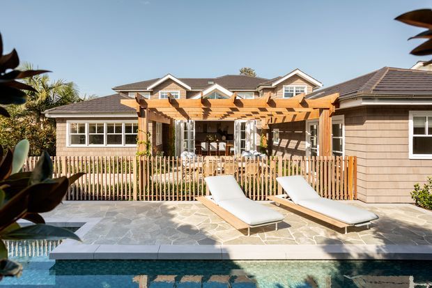 Sunny rear exterior of a low-rise residential home with a timber pergola-covered patio, outdoor dining area, pale horizontal cladding, pitched tiled roofs, and a swimming pool in the foreground. The pool terrace is paved in irregular light-grey stone and enclosed by a timber picket fence, creating a relaxed outdoor entertaining space.