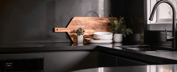 A dark, moody contemporary kitchen vignette with matte-black cabinetry and benchtops, a black splashback, and a timber serving board displayed against the wall. White bowls, potted greenery, and a tall black gooseneck tap provide contrast near the sink beneath a window.