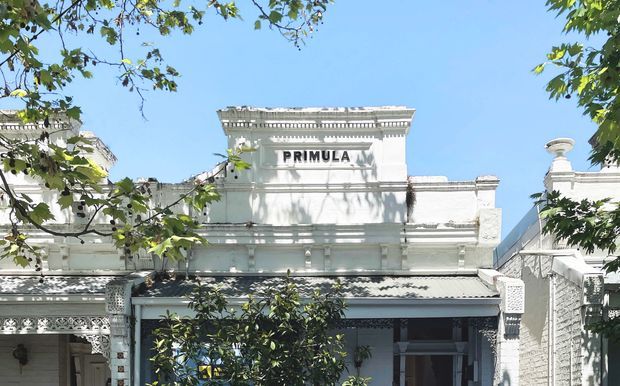 Front elevation of a historic white-painted building with ornate decorative mouldings, parapet detailing, a corrugated metal awning, and a prominent “PRIMULA” sign. The facade is partially framed by leafy tree branches under a clear blue sky.