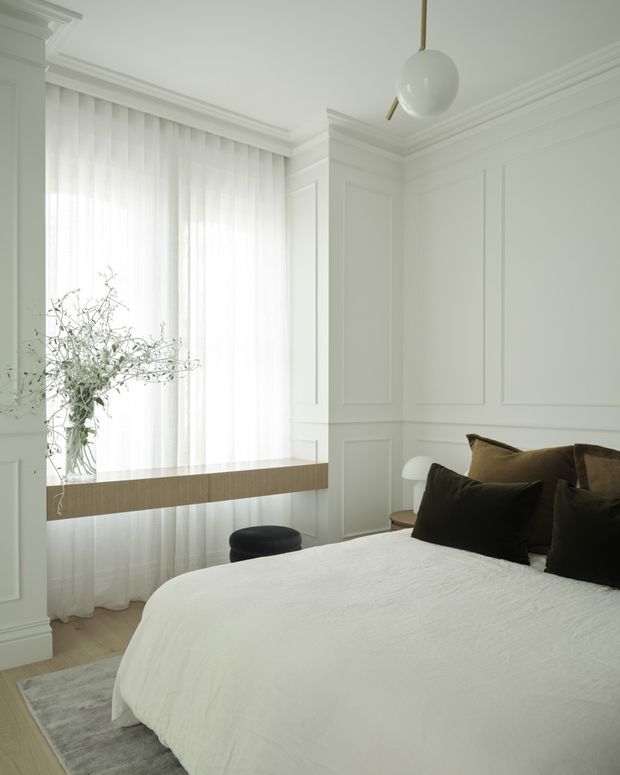 Bright, refined bedroom with white decorative wall panelling, ornate ceiling cornices, sheer floor-to-ceiling curtains, and a large upholstered bed dressed in white linen with dark cushions. A projecting timber-toned floating ledge and sculptural pendant light add contemporary contrast, while a small side table and organic branch arrangement complete the calm, monochromatic interior.