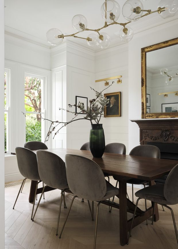 A refined dining room with a large dark timber dining table surrounded by upholstered dining chairs. White wall panelling, detailed cornice, tall windows, a decorative antique-style fireplace and ornate framed mirror create a classic architectural backdrop. A sculptural branch arrangement in a dark vase forms the centrepiece, while a contemporary brass-and-glass globe chandelier provides overhead lighting. The room is bright with a sophisticated blend of traditional architectural details and modern furnishings.