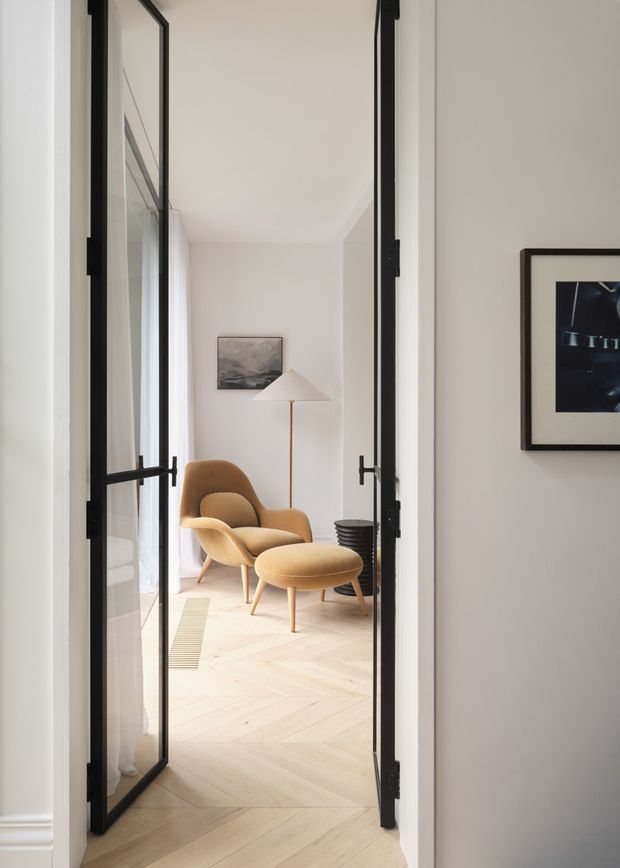 A bright, refined sitting area viewed through open black-framed glazed double doors. The room features pale herringbone timber flooring, soft off-white walls, a mustard upholstered lounge chair with matching ottoman, a sculptural floor lamp, a small dark side table, sheer curtains, and framed artwork. The composition highlights the connection between the hallway and the calm, minimalist seating space.