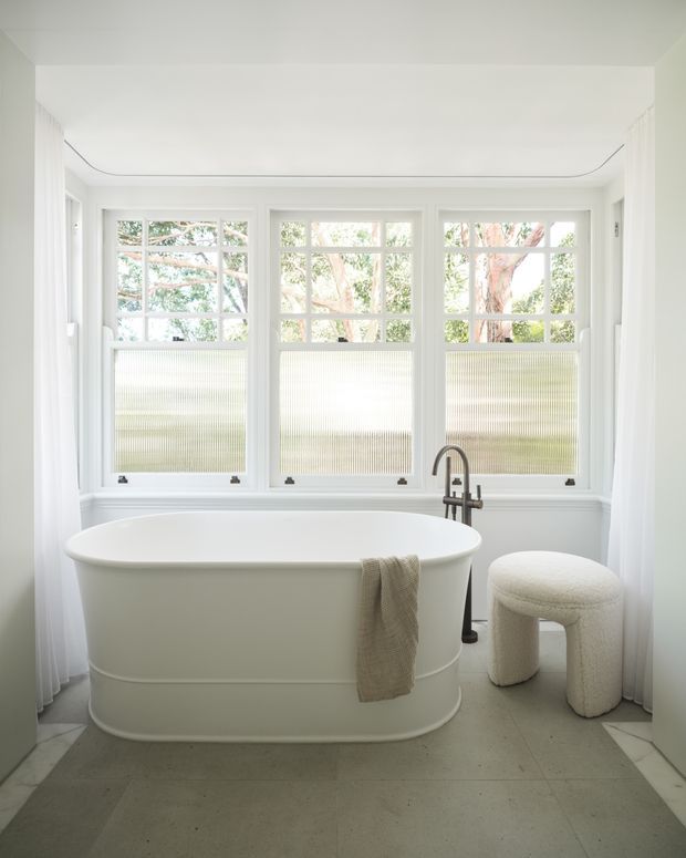Bright, refined bathroom centered on a freestanding oval bathtub beneath a large bank of white-framed windows with textured privacy glazing. A brushed metal floor-mounted tub filler, small upholstered stool, pale tiled floor, and sheer white curtains create a clean, airy, contemporary setting.