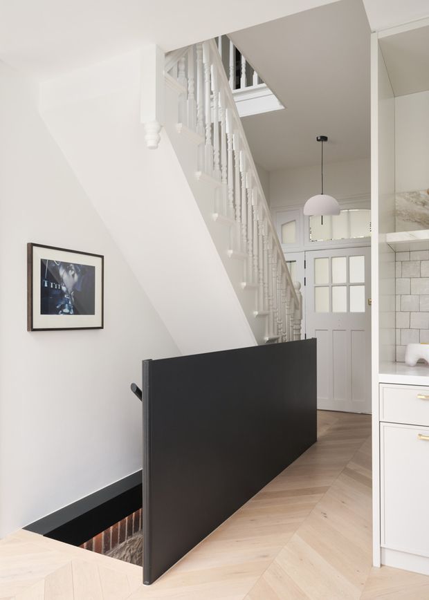 Bright, refined residential hallway and stairway with a white-painted traditional staircase, turned balusters, and a skylight above. A black steel-framed stairwell guard contrasts with the pale herringbone timber flooring and white walls. A paneled front door, framed artwork, pendant light, and part of a white kitchen with tiled splashback are visible.
