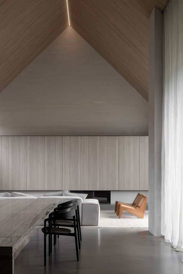 A refined open-plan interior with a dining area in the foreground and a living room beyond. The space features a dramatic pitched, timber-lined ceiling with a narrow linear skylight, full-height sheer curtains, pale vertical timber or wood-look wall cabinetry, a large stone dining table, dark dining chairs, a light sofa, and a sculptural timber lounge chair. The restrained neutral palette and clean detailing create a calm, minimalist atmosphere.