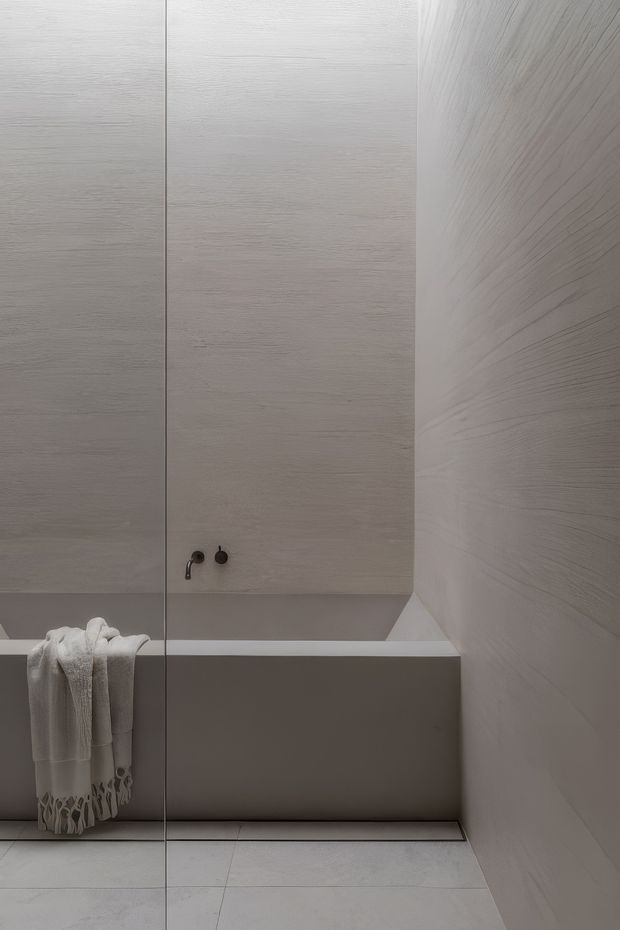 Minimalist contemporary bathroom featuring a built-in bathtub behind a frameless glass shower screen. The space is finished in pale textured wood-look wall panels and large light porcelain floor tiles, with dark wall-mounted bath controls and a draped white towel adding subtle contrast.
