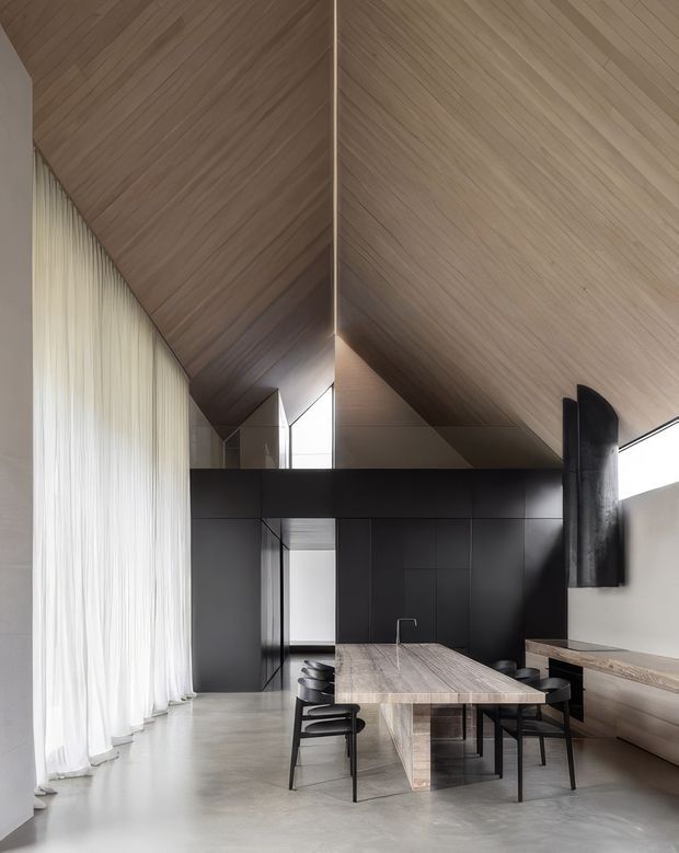A dramatic contemporary open-plan dining and kitchen space beneath a steeply angled timber-lined ceiling. A long stone or timber dining table with dark chairs occupies the foreground, while a minimalist black kitchen and island sit behind it. Full-height sheer curtains line the left wall, and a built-in bench runs along the right side beneath a narrow clerestory window. The palette is restrained, combining pale timber, concrete-grey flooring, matte black joinery, and abundant diffuse daylight.